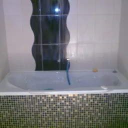 Biała wanna obudowana mozaiką w szaro-złote kwadraciki, na ścianie białe płytki z dekoracyjnym pionowym pasem z ciemnych płytek o falistym kształcie.